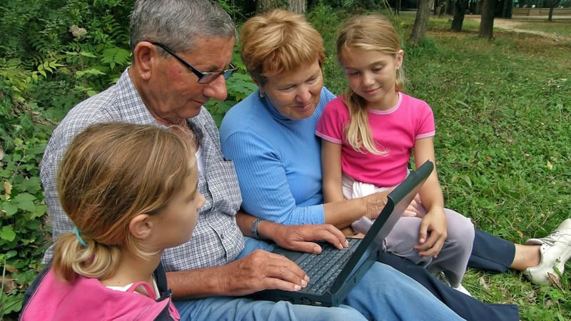 grandparents sitting on the ground outdoor with their granddaughters, all looking at a laptop computer, possibly even doing a puzzle