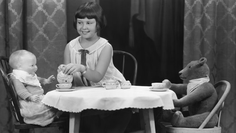 A little girl having a tea party with her doll and teddy bear. An essayist discusses childhood games, then the games people play as adults.
