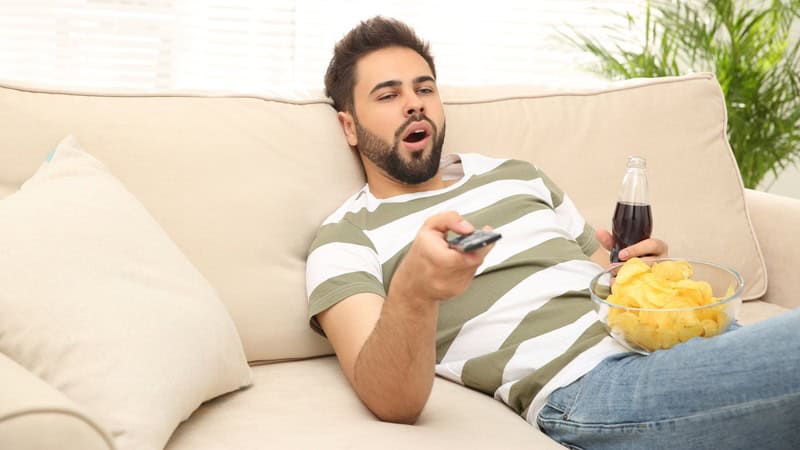 A lazy young adult male yawning, slouching on the sofa with a soda, chips, and TV remote, like the sons in this article who are freeloading off of their unappreciated, overworked mom. Image by Chernetskaya.