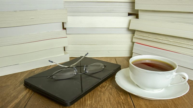 Three stacks of books behind an e-reader, reading glasses, and cup of tea or coffee. By Karen Foley. For essay, making time for books