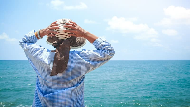 Woman holding her hat as she glances over a rich turquoise-blue ocean. Safe Solo Travel for Older Women. (Dreamstime/TCA)